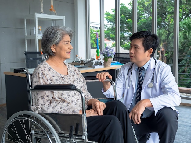 Souriante heureuse patiente âgée asiatique âgée assise en fauteuil roulant parlant avec un homme sympathique médecin en blouse blanche dans un cabinet médical à l'hôpital Soins de santé et concept médical