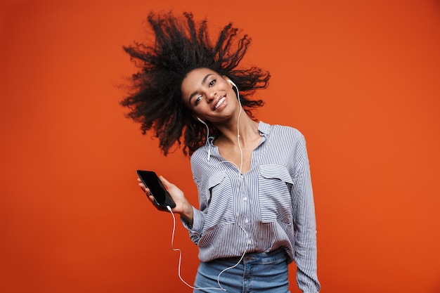 Souriante jeune belle femme africaine portant des vêtements décontractés debout isolée sur un mur rouge, écoutant de la musique avec des écouteurs et un téléphone portable, dansant