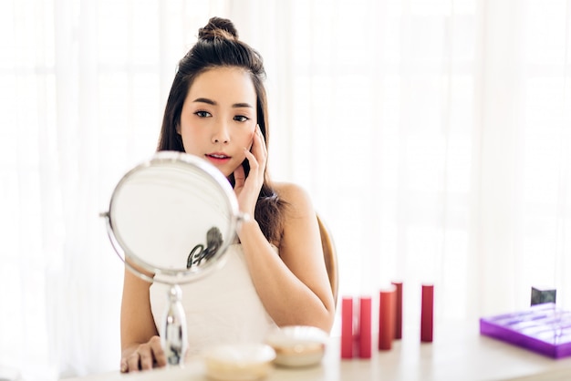 Souriante jeune belle femme asiatique fraîche peau saine regardant sur miroir avec des cosmétiques de maquillage mis à la maison