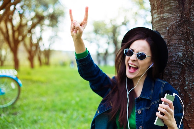 Souriante jeune belle fille hipster écoute de la musique rock du téléphone et s'amuse sous elle à l'extérieur