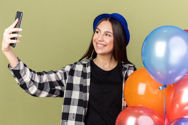 Souriante jeune belle fille portant un chapeau bleu debout à proximité des ballons et prendre un selfie