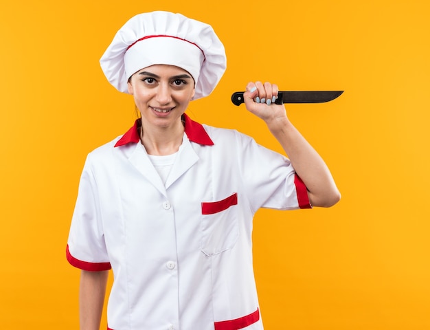 Souriante jeune belle fille en uniforme de chef tenant un couteau isolé sur un mur orange