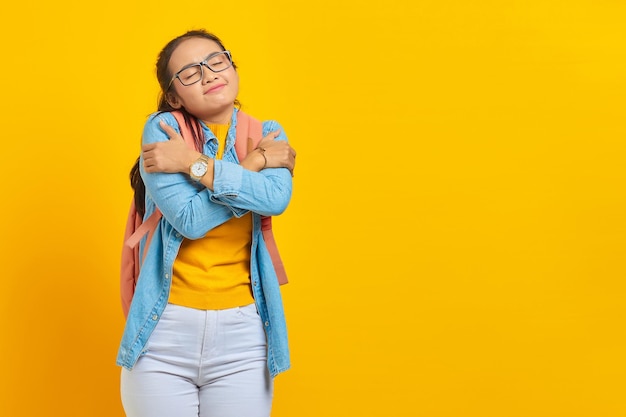 Souriante jeune étudiante asiatique en tenue denim avec sac à dos se serrant dans ses bras et se sentant froide isolée sur fond jaune Éducation au concept universitaire