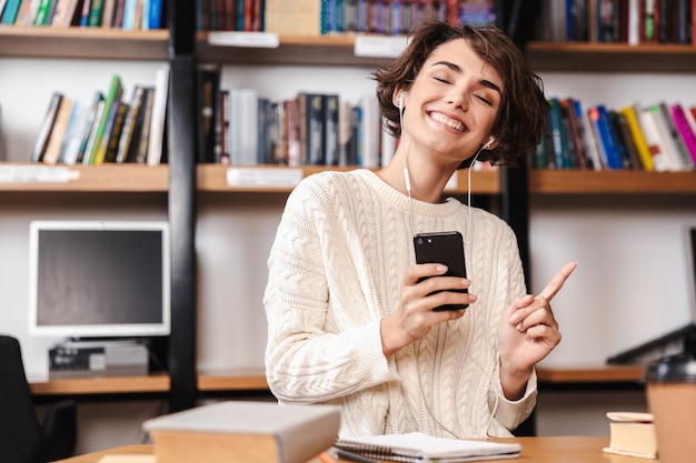 Souriante jeune étudiante étudiant assis au bureau de la bibliothèque, tenant un téléphone mobile