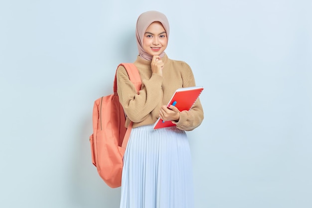 Souriante jeune étudiante musulmane asiatique en pull marron avec sac à dos tenant un livre et pensant à quelque chose d'isolé sur fond blanc retour au concept d'école