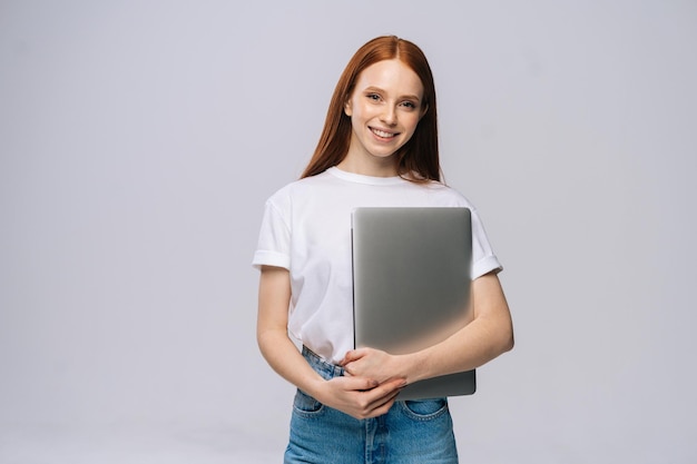 Souriante jeune étudiante tenant un ordinateur portable et regardant la caméra sur fond isolé