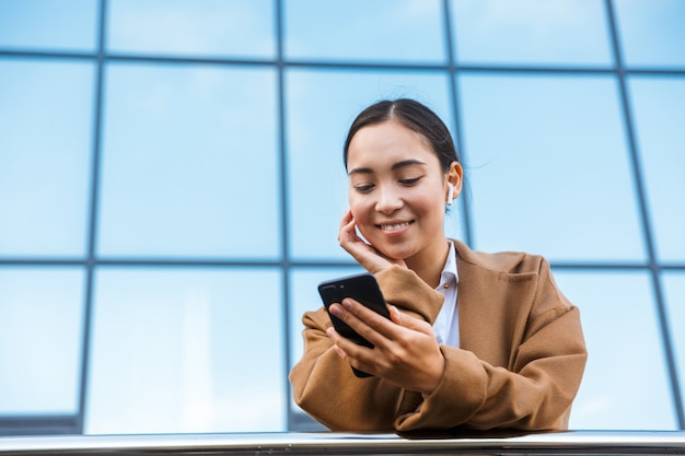 Souriante jeune femme d'affaires asiatique portant un manteau debout devant un bâtiment en verre à l'extérieur de la ville, à l'aide d'un téléphone portable