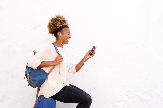 Souriante jeune femme africaine assise sur une valise et regardant le téléphone