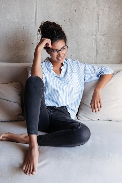 Souriante jeune femme africaine portant une chemise relaxante sur un canapé à la maison