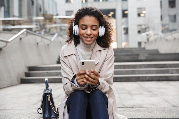 Souriante jeune femme africaine portant un manteau d'automne assise sur un escalier dans la rue, utilisant un téléphone portable tout en écoutant de la musique avec des écouteurs sans fil