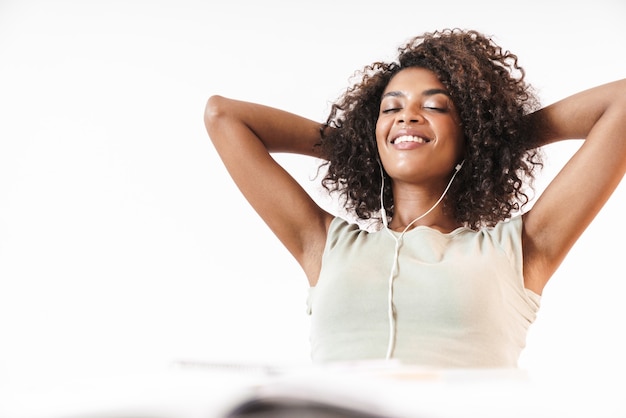Souriante jeune femme africaine se relaxant tout en étant assise au bureau isolé sur un mur blanc, écoutant de la musique avec des écouteurs