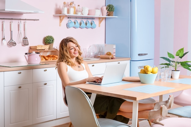 Souriante jeune femme à l'aide d'un ordinateur portable dans la cuisine à la maison. Femme blonde travaille sur ordinateur, pigiste ou blogueur travaillant à la maison
