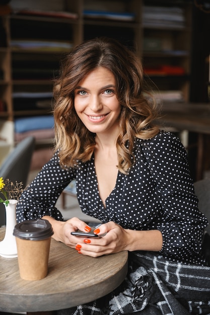 Souriante jeune femme à l'aide de téléphone portable assis au café, boire du café