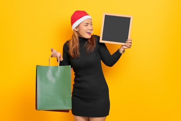 Souriante jeune femme asiatique en chapeau de père Noël tenant des sacs à provisions et tableau isolé sur fond jaune