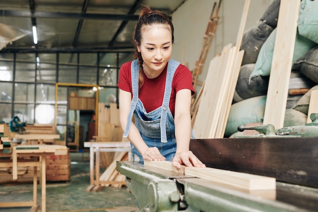 Souriante jeune femme asiatique charpentier coupant une planche de bois en planches