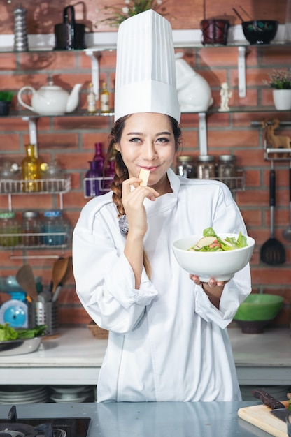 Souriante jeune femme asiatique chef cuisinier en uniforme blanc debout à la cuisine, montrant la salade dans un bol et pomme rouge sur sa main.