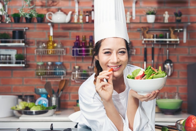 Souriante jeune femme asiatique chef cuisinier en uniforme blanc debout à la cuisine, montrant la salade dans un bol et pomme rouge sur sa main.