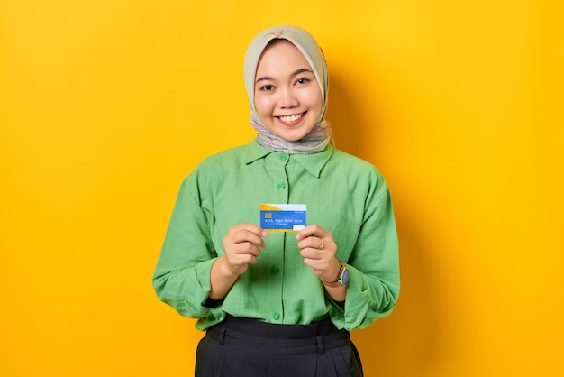 Souriante jeune femme asiatique dans une chemise verte montrant une carte de crédit sur fond jaune