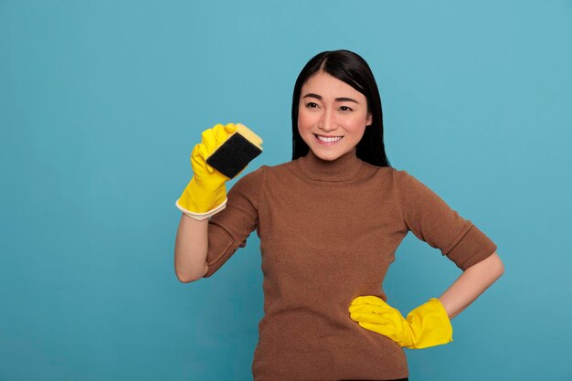 Souriante jeune femme asiatique heureuse et joyeuse tenant une éponge avec des gants jaunes isolés sur fond bleu, femme au foyer, concept de maison de nettoyage, rire joyeux et état d'esprit positif