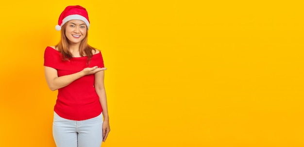 Souriante jeune femme asiatique portant un chapeau de Noël et tendant la main sur le côté invitant à venir