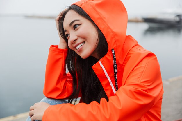 Souriante jeune femme asiatique portant un imperméable passer du temps à l'extérieur à marcher sur la côte