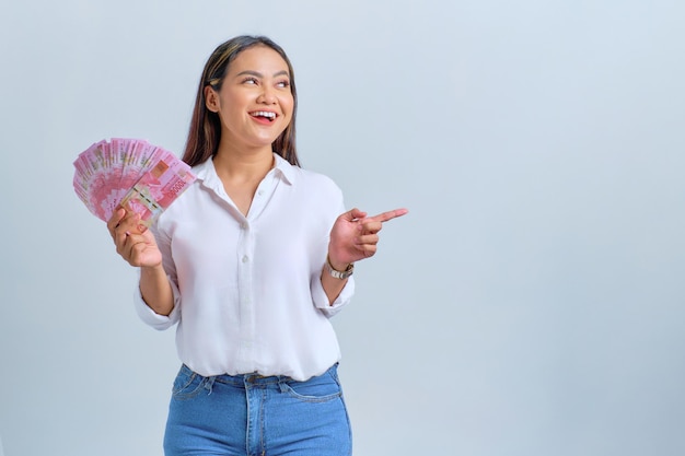 Souriante jeune femme asiatique tenant des billets de banque et pointant du doigt l'espace de copie isolé sur fond blanc