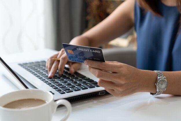 Souriante jeune femme asiatique tenant une carte de crédit tout en utilisant un ordinateur portable au bureau à domicile