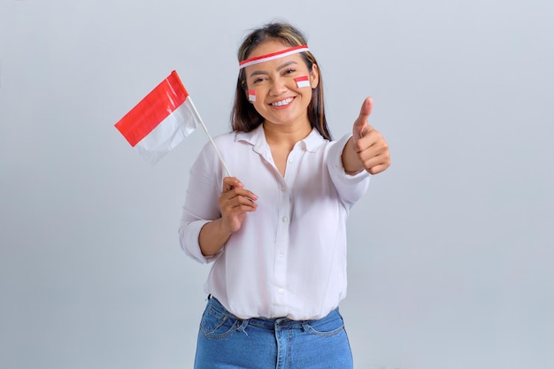 Souriante jeune femme asiatique tenant le drapeau indonésien tout en montrant les pouces vers le haut isolé sur fond blanc Concept de la fête de l'indépendance indonésienne