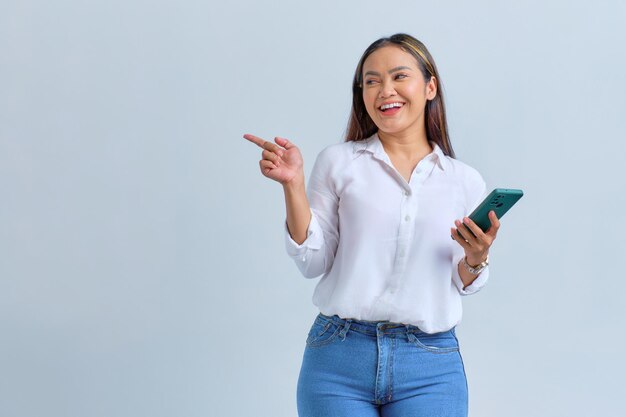 Souriante jeune femme asiatique tenant un téléphone portable et pointant du doigt l'espace de copie isolé sur fond blanc