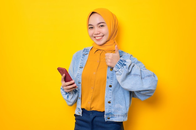 Souriante jeune femme asiatique en veste de jeans tenant un téléphone portable et faisant le geste du pouce vers le haut approuver une bonne chose isolée sur fond jaune