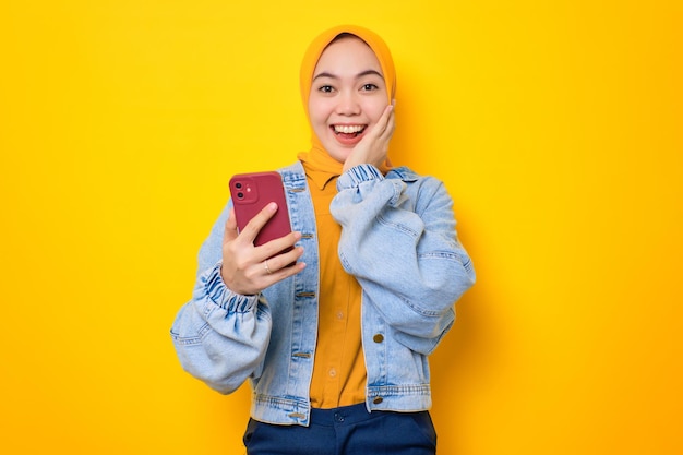 Souriante jeune femme asiatique en veste de jeans tenant un téléphone portable et regardant la caméra isolée sur fond jaune