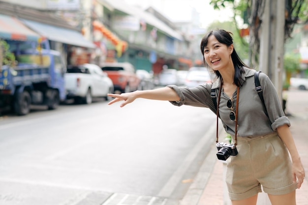 Souriante jeune femme asiatique voyageur faisant de l'auto-stop sur une route de la ville La vie est un concept de voyage