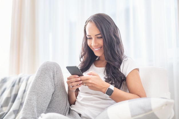 Souriante jeune femme assise sur un canapé dans un salon souriant au téléphone passant du temps en ligne pendant un verrouillage.