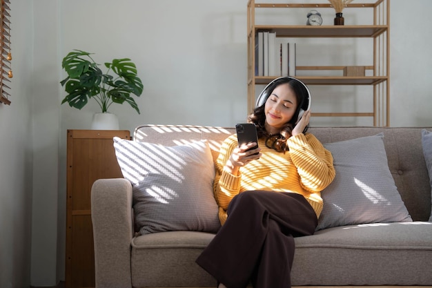 Souriante jeune femme assise sur un canapé à la maison parler lors d'un appel vidéo sur un smartphone avec un casque