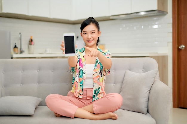 Souriante jeune femme assise sur le canapé à la maison, pointant l'index sur le téléphone mobile