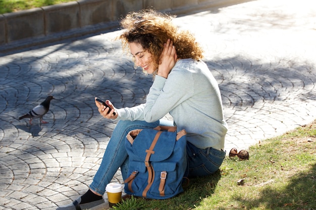 Souriante jeune femme assise dans un parc à la recherche de téléphone portable