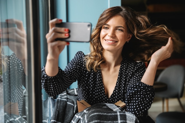 Souriante jeune femme assise enveloppée dans une couverture à la fenêtre, prenant un selfie