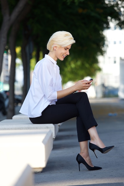 Souriante jeune femme assise à l&#39;extérieur de lire un message texte