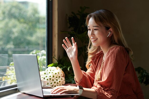 Souriante jeune femme assise à la fenêtre d'un café et d'un collègue d'appel vidéo pour discuter des détails du travail et du projet