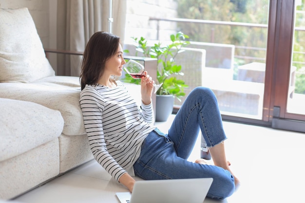 Souriante jeune femme assise sur le sol avec un ordinateur portable et discutant avec des amis buvant du vin