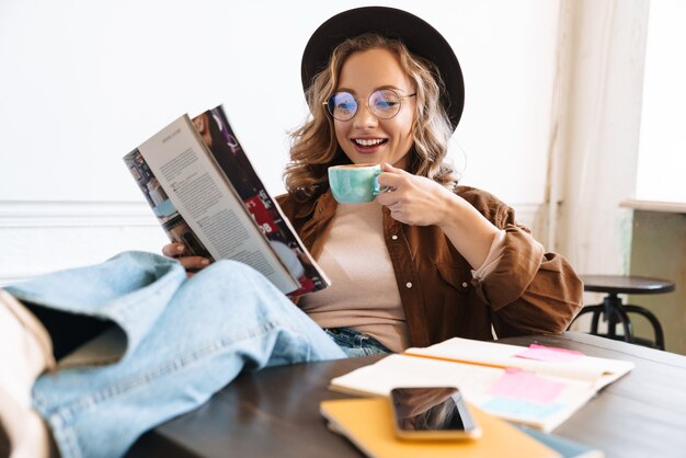 Souriante jeune femme au chapeau lisant un magazine avec les jambes sur la table en buvant du café