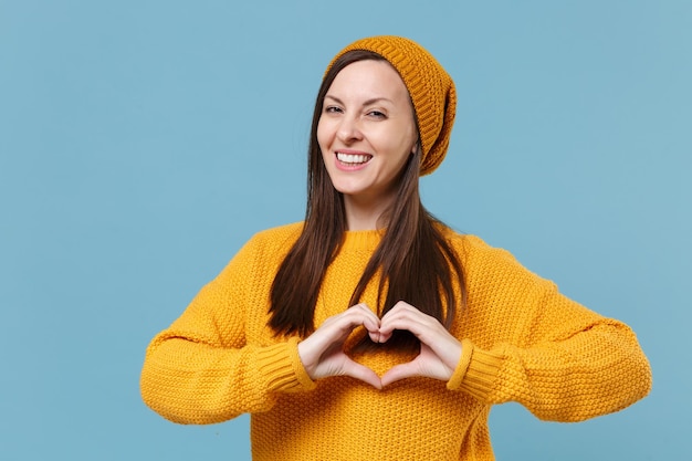 Souriante jeune femme au chapeau pull jaune posant isolé sur fond bleu portrait en studio. Concept de mode de vie des émotions des gens. Maquette de l'espace de copie. Montrant le coeur en forme avec les mains, signe en forme de coeur.