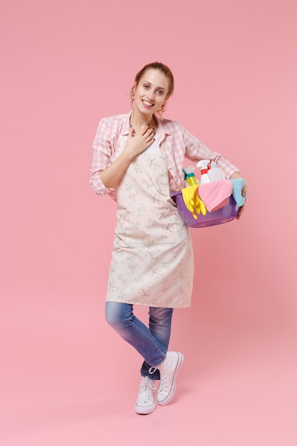 Souriante jeune femme au foyer dans un bassin de maintien de tablier avec des bouteilles de détergent lavant des nettoyants faisant des travaux ménagers isolés sur un portrait de studio de fond de mur rose. Notion d'entretien ménager. Mettez la main sur la poitrine.