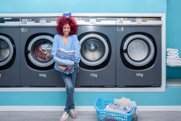 Souriante jeune femme aux cheveux afro rouges tenant des vêtements dans une laverie automatique bleue