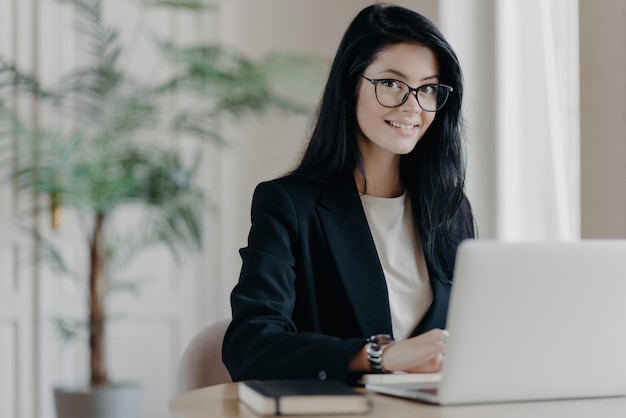 Souriante jeune femme bien habillée secrétaire aux cheveux noirs travaille sur un ordinateur portable au bureau rend le travail de projet ou de recherche Directeur des ventes dans un bureau moderne lieu de travail confortable Concept de travail à distance