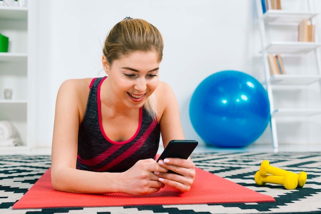 Souriante jeune femme blonde allongée sur un tapis d&#39;exercice rouge à l&#39;aide d&#39;un téléphone portable