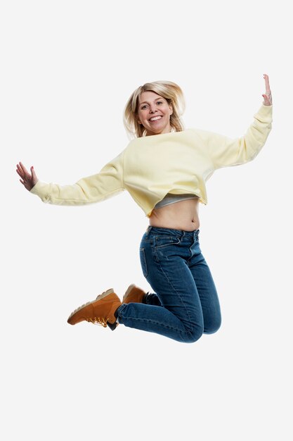 Souriante jeune femme blonde en jeans et un pull jaune saute. Activité et positivité. Isolé sur fond blanc. Verticale.