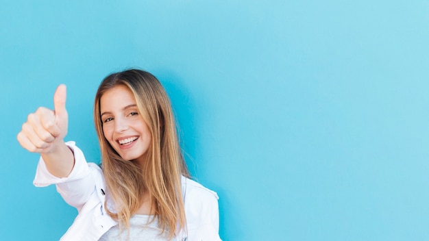 Souriante jeune femme blonde montrant le pouce en haut signe sur fond bleu