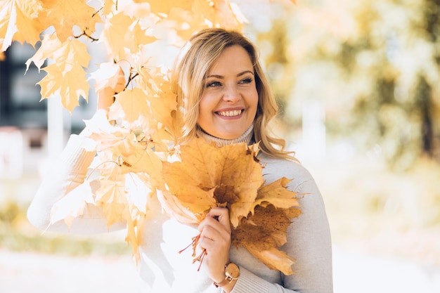 Souriante jeune femme blonde vêtue de beige debout à l'extérieur peut-être dans le parc d'automne avec un tas de feuilles dans ses mains. Fond d'automne jaune et orange coloré avec effet flou