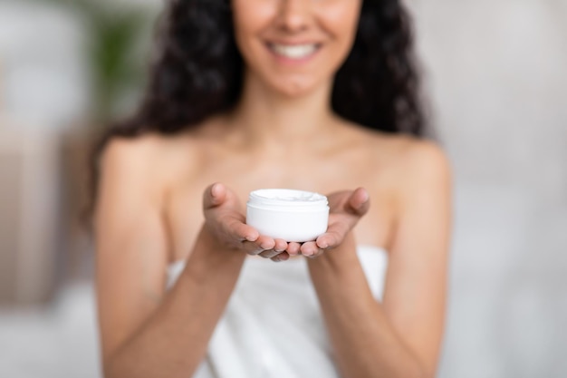Photo souriante jeune femme brune caucasienne aux cheveux longs dans une serviette spectacle pot de crème dans la chambre